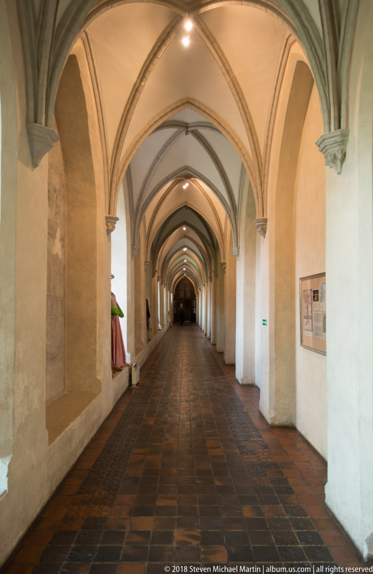 Long pale corridor with repeated pointed rib vaults, narrow wall openings, and a dark tiled floor receding into the distance.