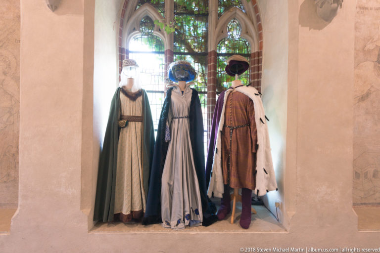 Three mannequins in medieval-style gowns, cloaks, fur trim, and headwear displayed in a tall Gothic window recess.