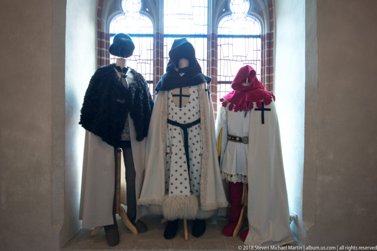 Three mannequins in medieval-style military dress and pale cloaks bearing dark crosses, posed before a tall window.