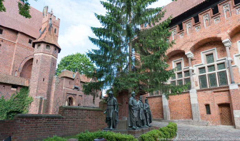 Bronze historical figures stand in a small landscaped courtyard beside ornate red-brick castle walls, arched windows, ivy, and trees.