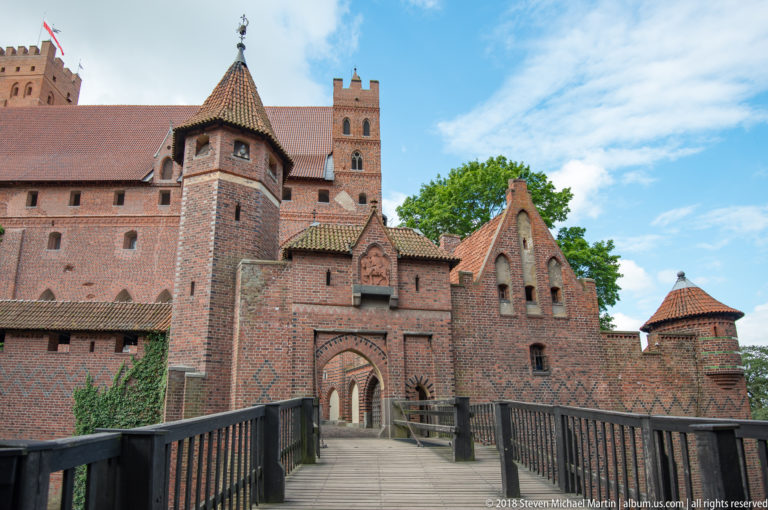 Wooden bridge with dark railings leading toward a decorative brick gatehouse, towers, and taller castle buildings beyond.