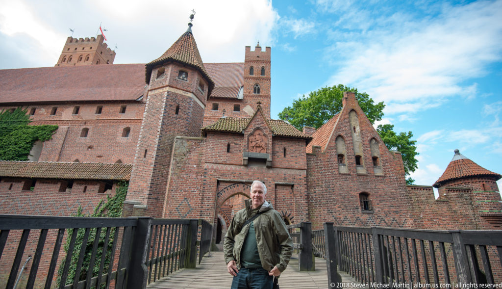 Visitor standing on a wooden castle bridge before a decorative red-brick gatehouse, towers, and steep tiled roofs.