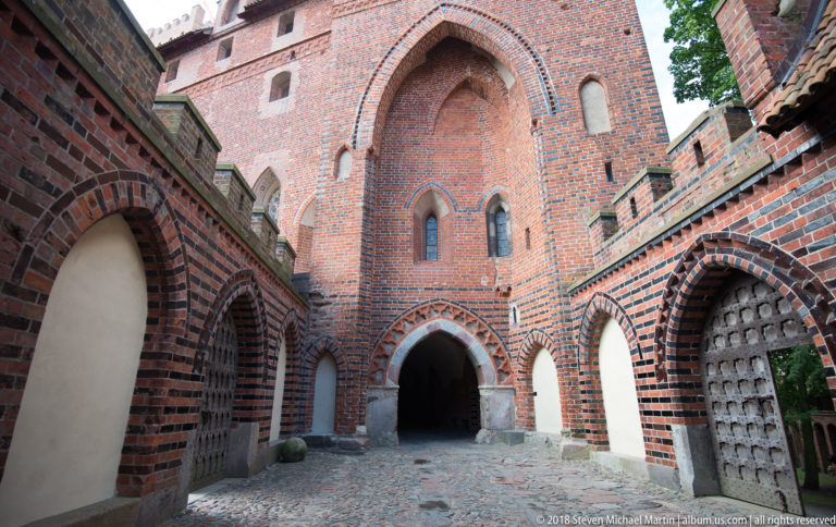 Cobbled brick court with a tall recessed pointed arch, smaller side arches, patterned masonry, and heavy timber doors.