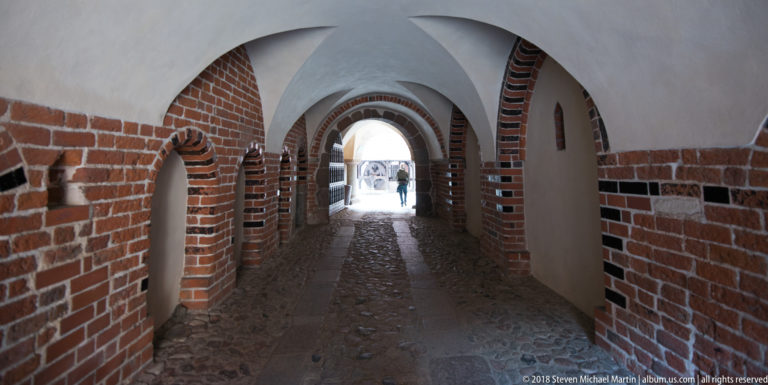 Narrow vaulted passage with repeated brick-and-plaster arches, small wall openings, uneven stone paving, and bright daylight at the far end.