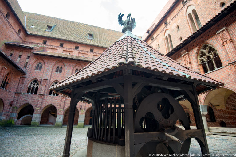 Roofed timber well or hoisting structure with large wheels and a bird finial, standing in a Gothic brick courtyard.