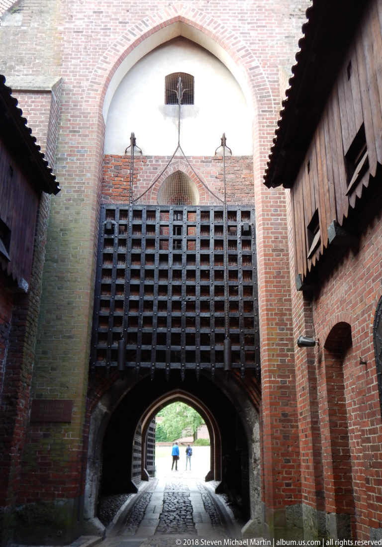 Heavy timber lattice gate beneath a pointed brick arch, seen from inside a narrow fortified passage with wooden side structures.