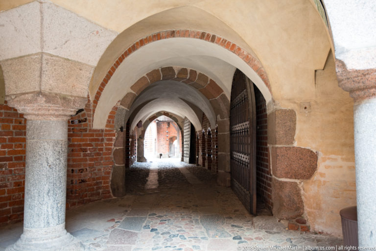 Vaulted castle passage with brick arches, a cylindrical stone column, thick masonry piers, an iron gate, and stone paving.