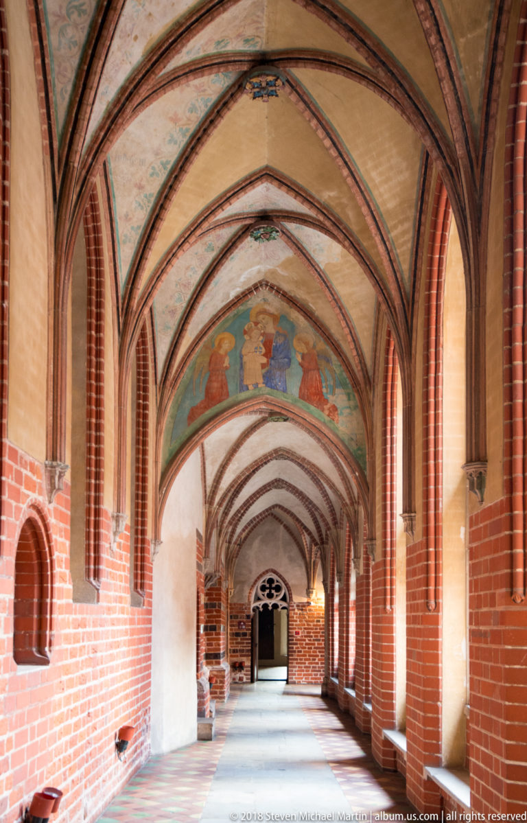 Long Gothic gallery with red-brick rib vaults, painted decorative vault panels, and daylight entering through openings along one side.