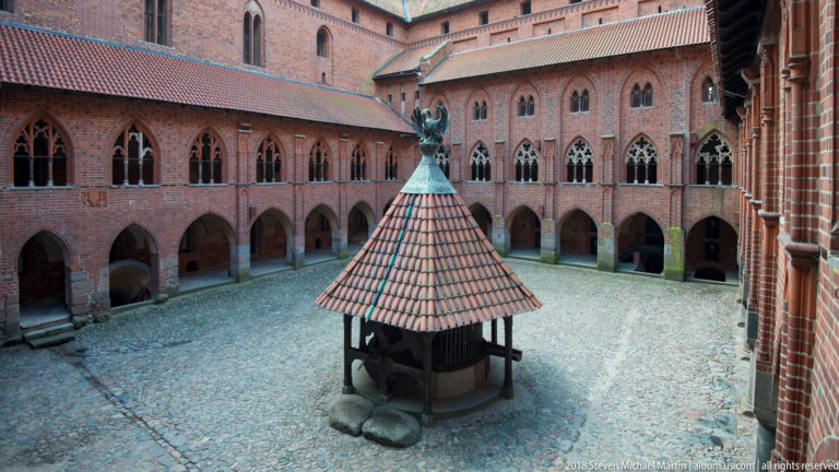 Elevated view of a square Gothic brick courtyard with two levels of pointed arcades and a roofed timber well structure below.