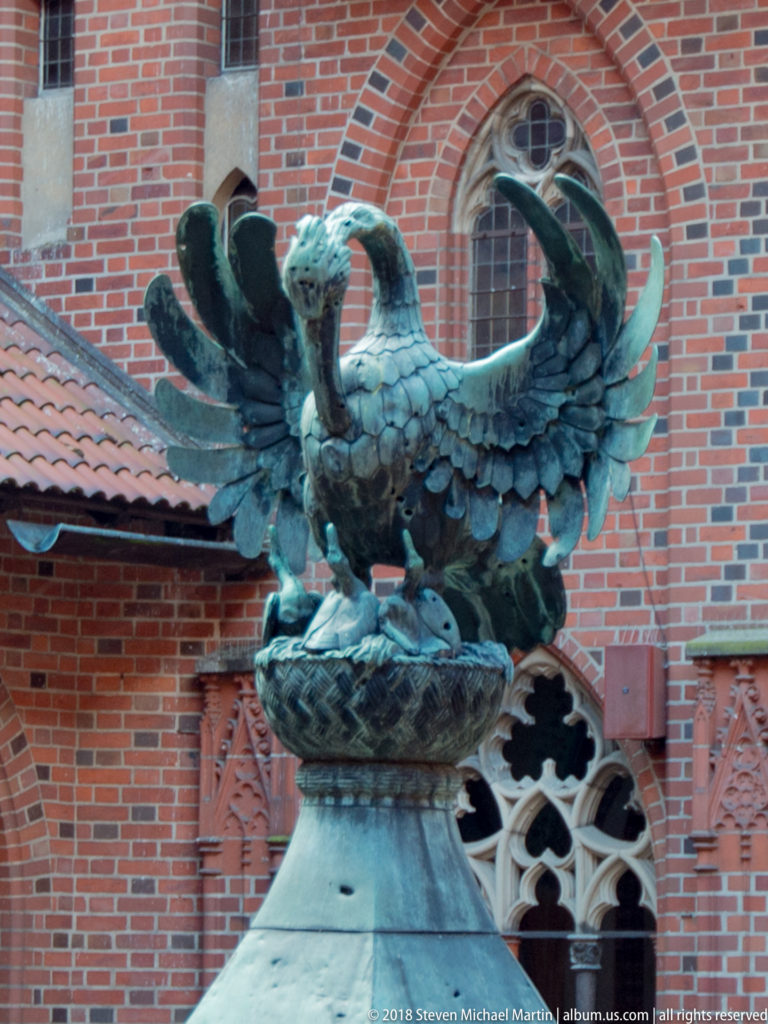 Close view of a dark metal bird sculpture with spread wings against red-brick Gothic windows and decorative masonry.