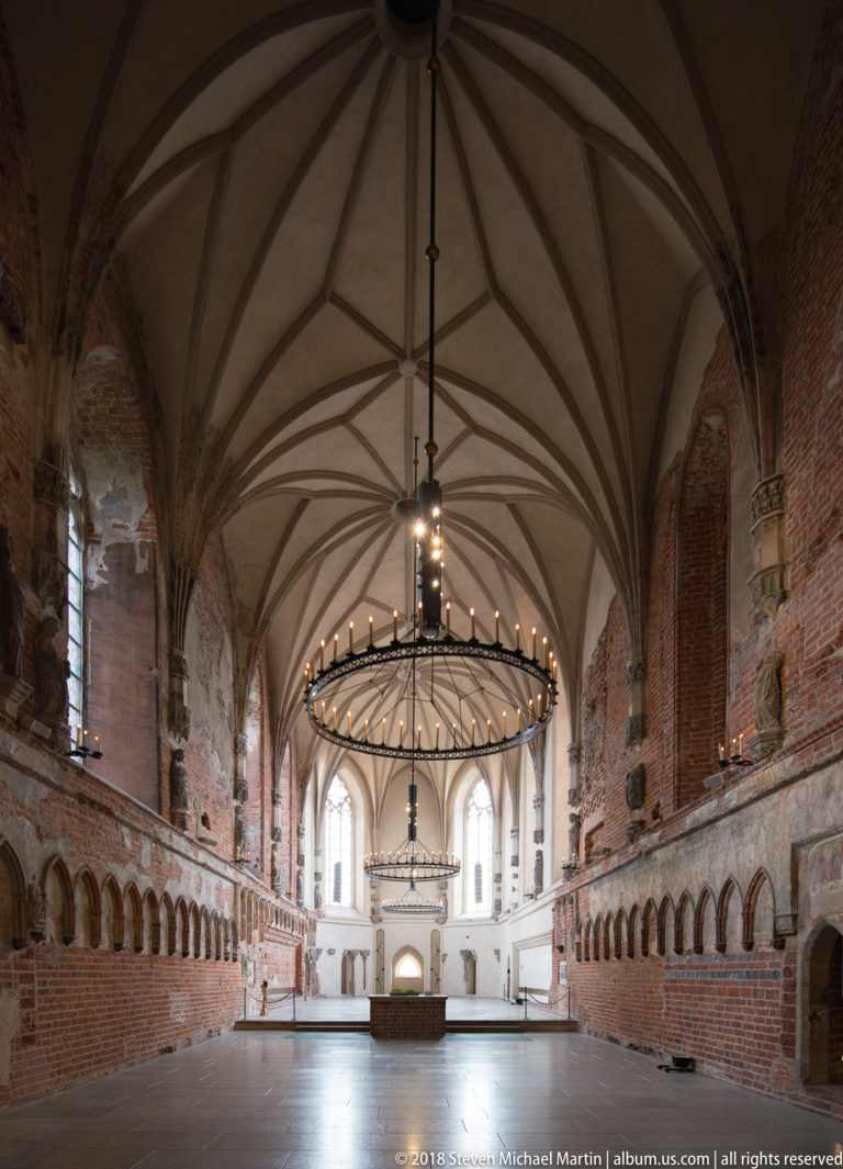Large brick hall with geometric rib vaulting, tall windows, circular chandeliers, and a low platform at the far end.