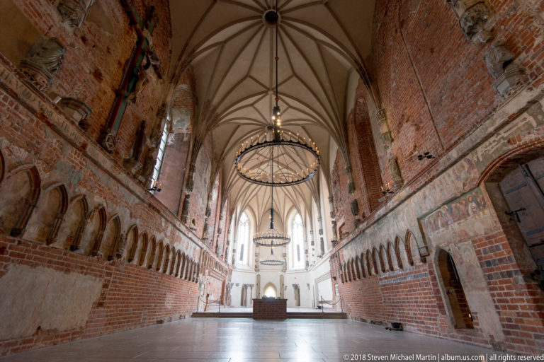 Axial interior view of a long brick hall with ribbed vaulting, wall niches, circular chandeliers, and a bright far end.