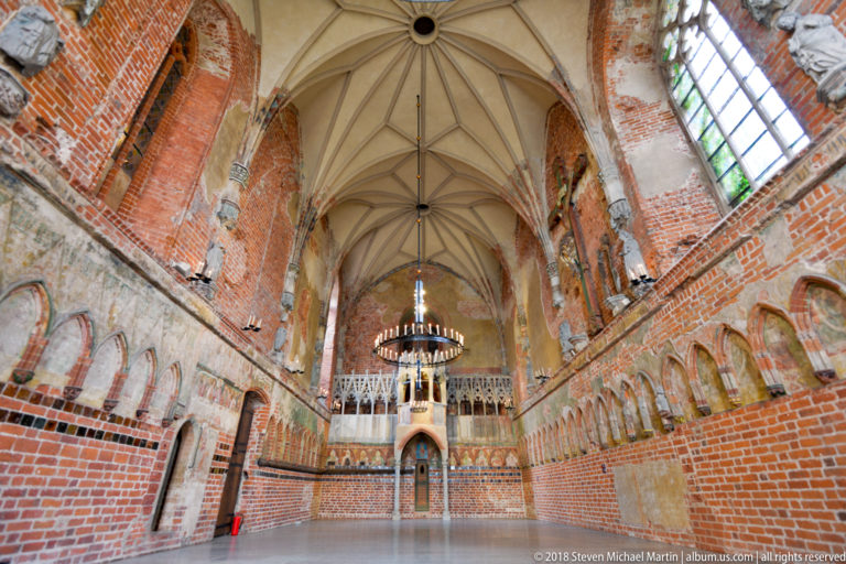 Large vaulted brick hall with circular chandeliers and a raised stone gallery or screen across the far wall.