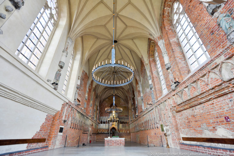 High vaulted hall with large windows, pale restored walls, exposed red brick, circular chandeliers, and a raised gallery in the distance.