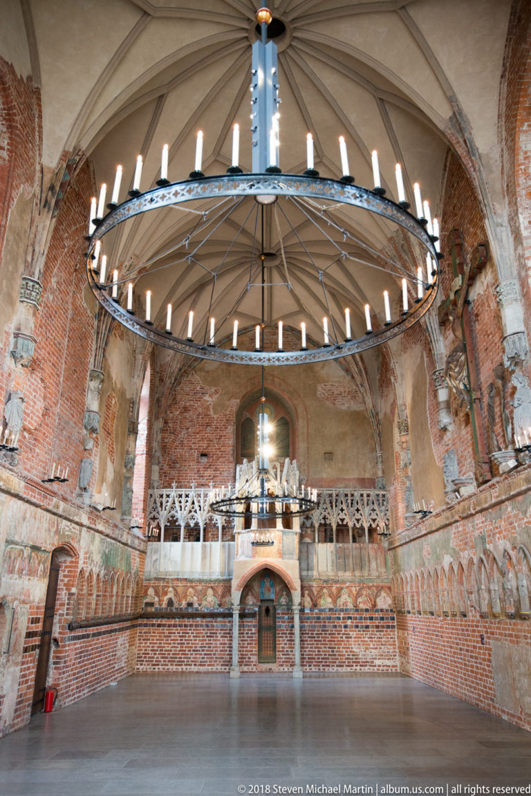 Large circular chandelier suspended in front of a pale stone gallery, with brick walls, rib vaulting, and traces of painted decoration.