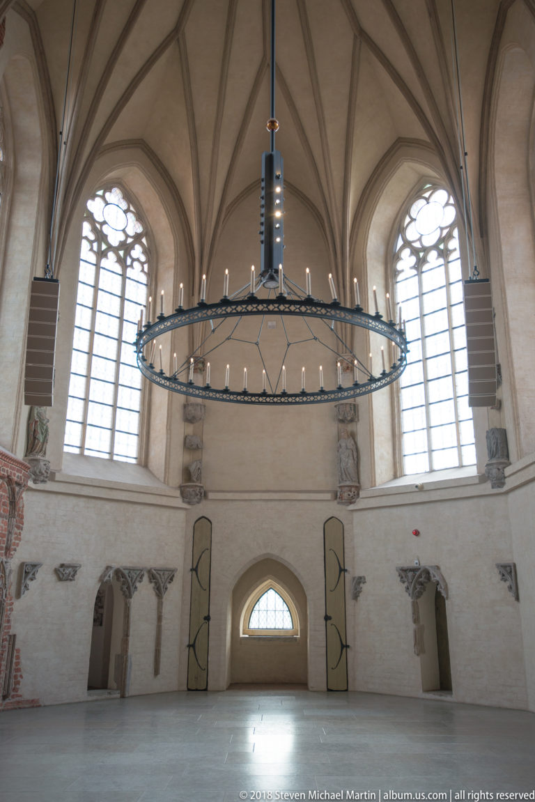 Large circular chandelier suspended before tall Gothic windows in a pale vaulted room with small doorways and architectural fragments.