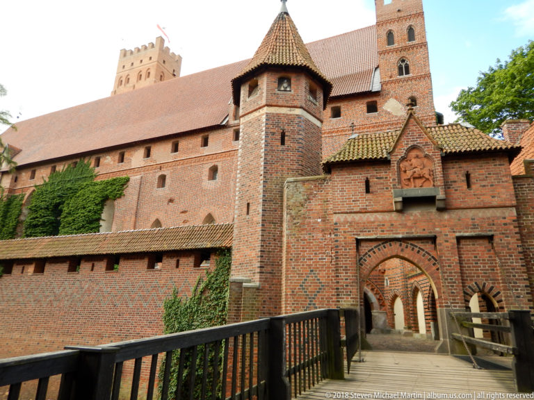 Bridge approaching a pointed brick gateway beneath a square tower, with repeated arches and a small relief above the entrance.