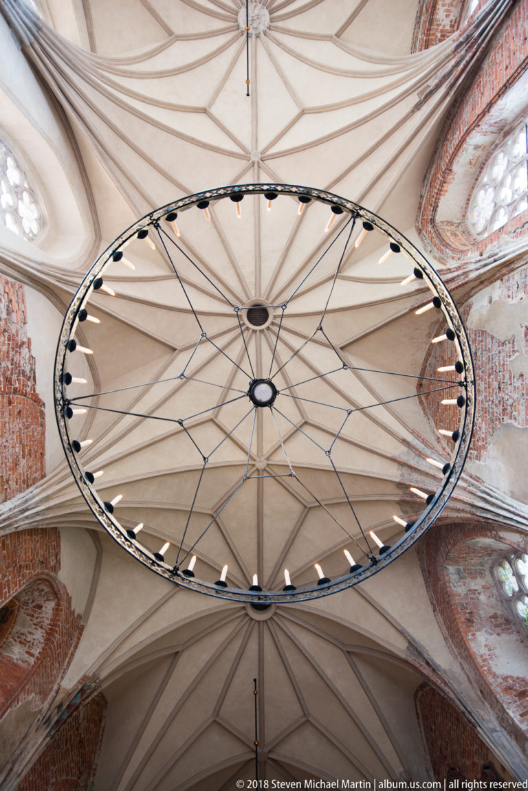 Large circular ring chandelier suspended beneath a pale rib-vaulted ceiling, with red brick and Gothic windows at the edges.