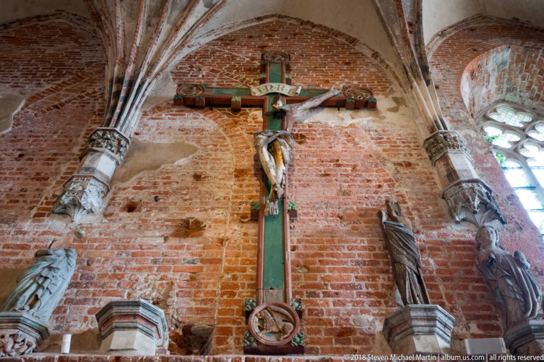 Large painted crucifix with Christ mounted on an exposed brick wall, flanked by carved religious figures beneath Gothic vaulting.