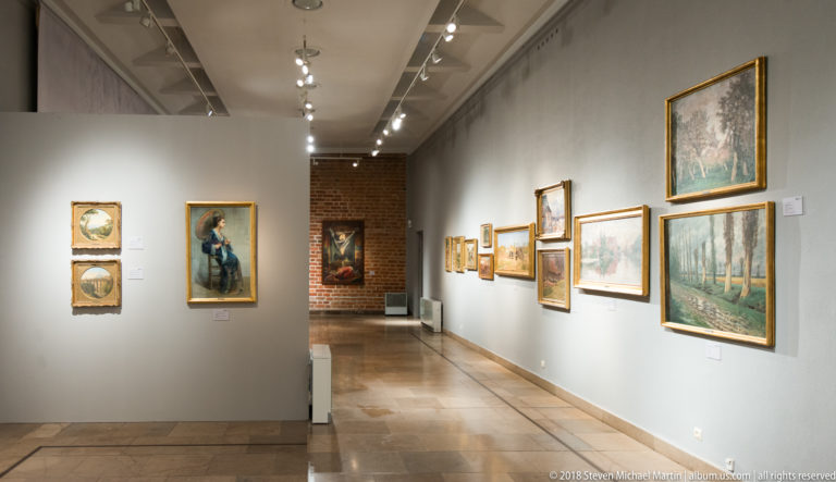 Interior museum gallery with framed portraits and landscapes on pale walls, track lighting overhead, and exposed brick at the far end.