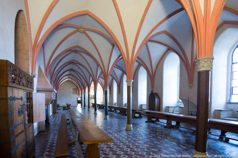 Long Gothic hall with red rib vaults, slender columns, wooden tables and benches, patterned floor tiles, and tall windows.