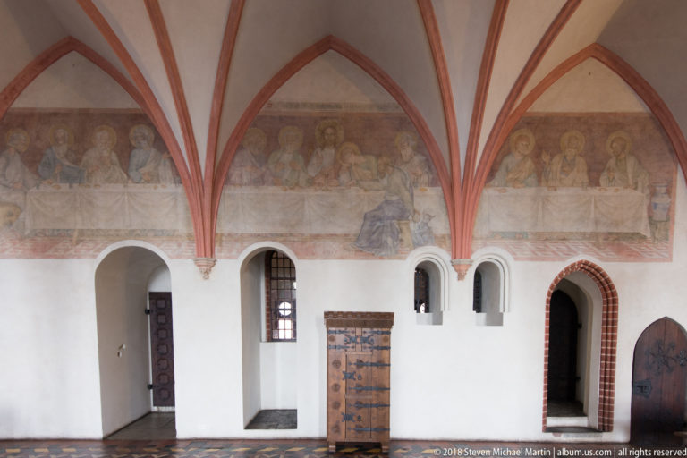 Faded mural of seated sacred figures above arched doorways and an iron-bound wooden chest in a vaulted castle interior.