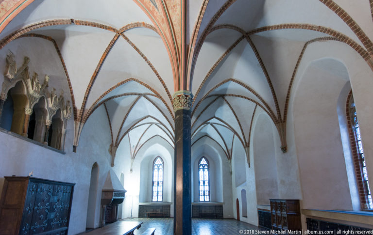 Vaulted Gothic chamber with a central dark column, radiating red ribs, tall pointed windows, wall niches, and wooden furnishings.