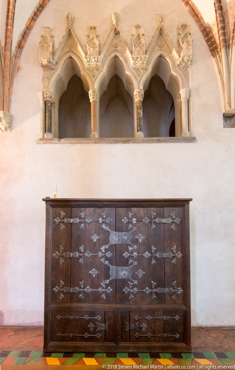 Triple cusped Gothic stone niche with slender columns above a large dark wooden chest decorated with repeated metal fittings.