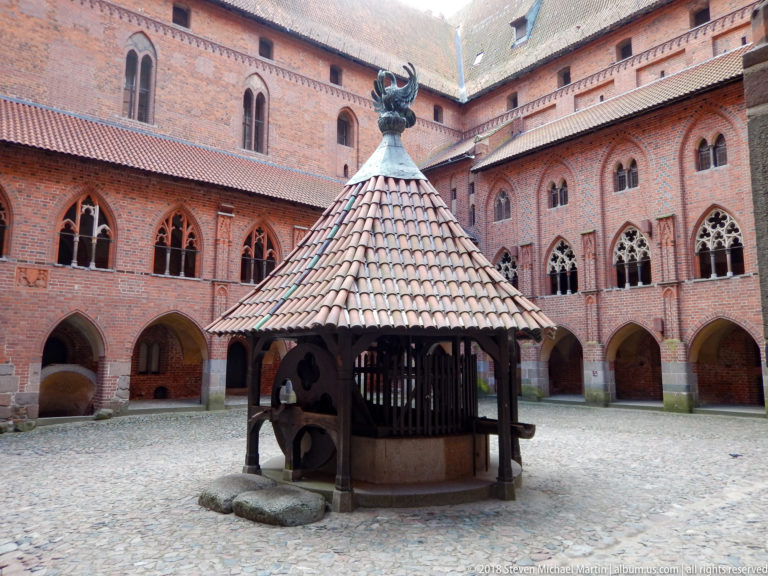 Roofed timber well in the center of a cobbled courtyard surrounded by red-brick Gothic arcades and pointed windows.