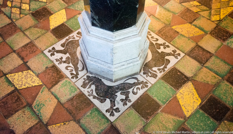 Multicolored medieval-style floor tiles and a pale heraldic border surrounding the octagonal stone base of a dark column.