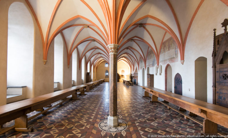 Long Gothic hall with central columns, red rib vaults, patterned tile floor, wooden benches, and arched doorways.