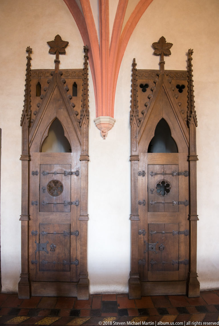 Two tall iron-bound wooden doors in ornate pointed Gothic stone surrounds with crockets, finials, and red vault ribs above.