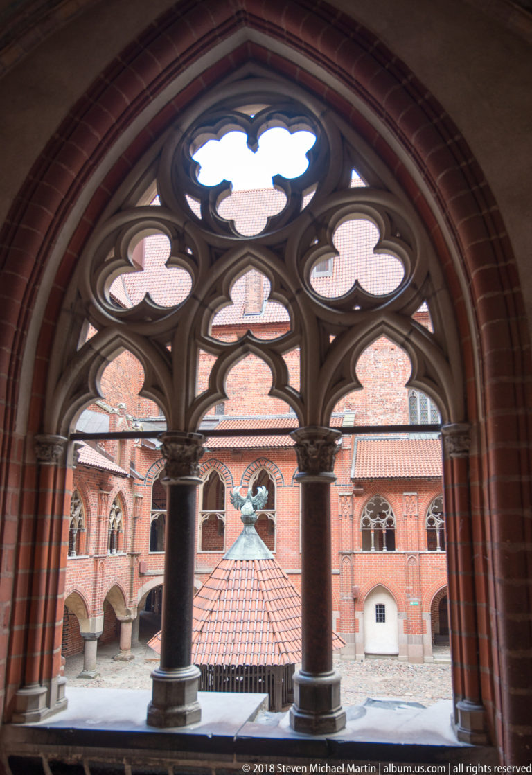 Large pointed Gothic tracery window framing red-brick courtyard buildings, arcades, and tiled roofs beyond.