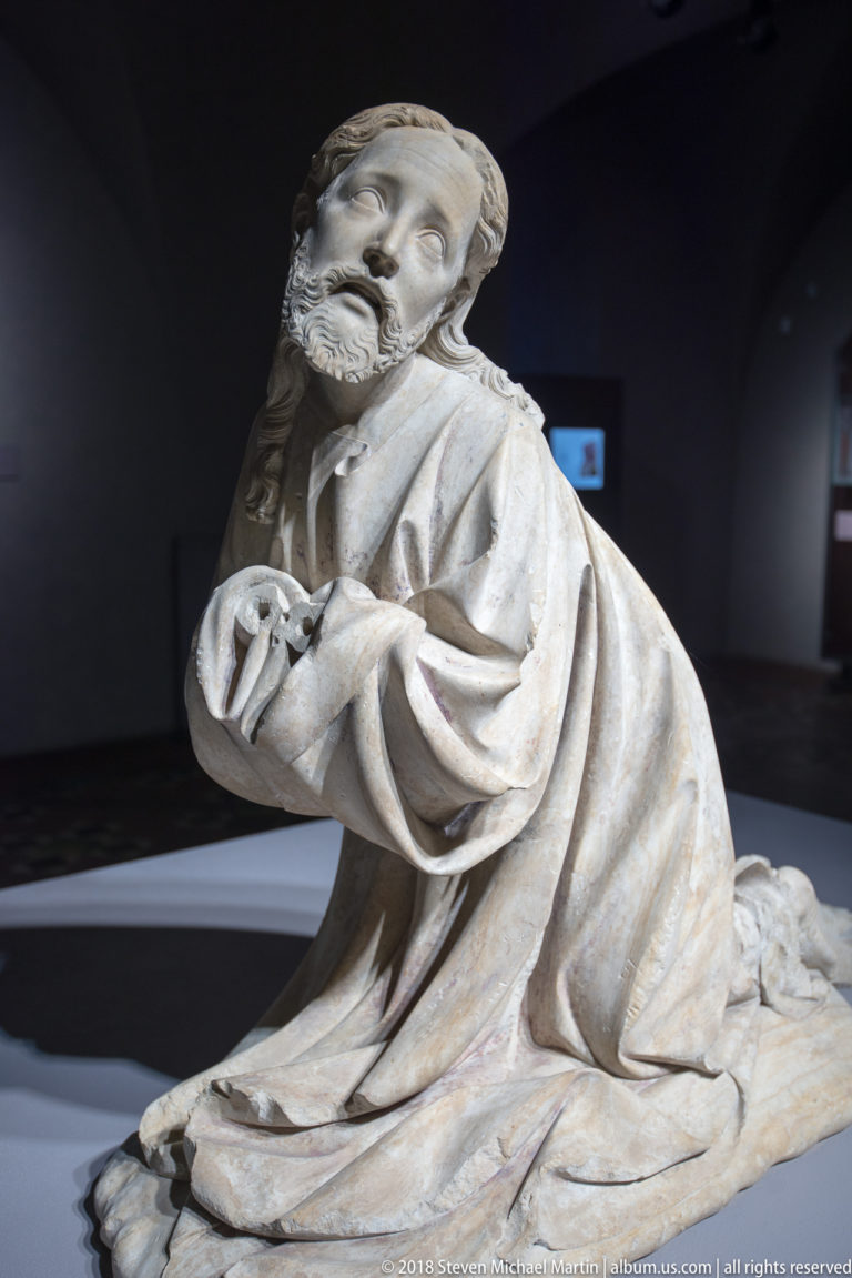 Carved pale stone figure of a bearded man kneeling with clasped hands, draped in heavy robes against a dark museum background.