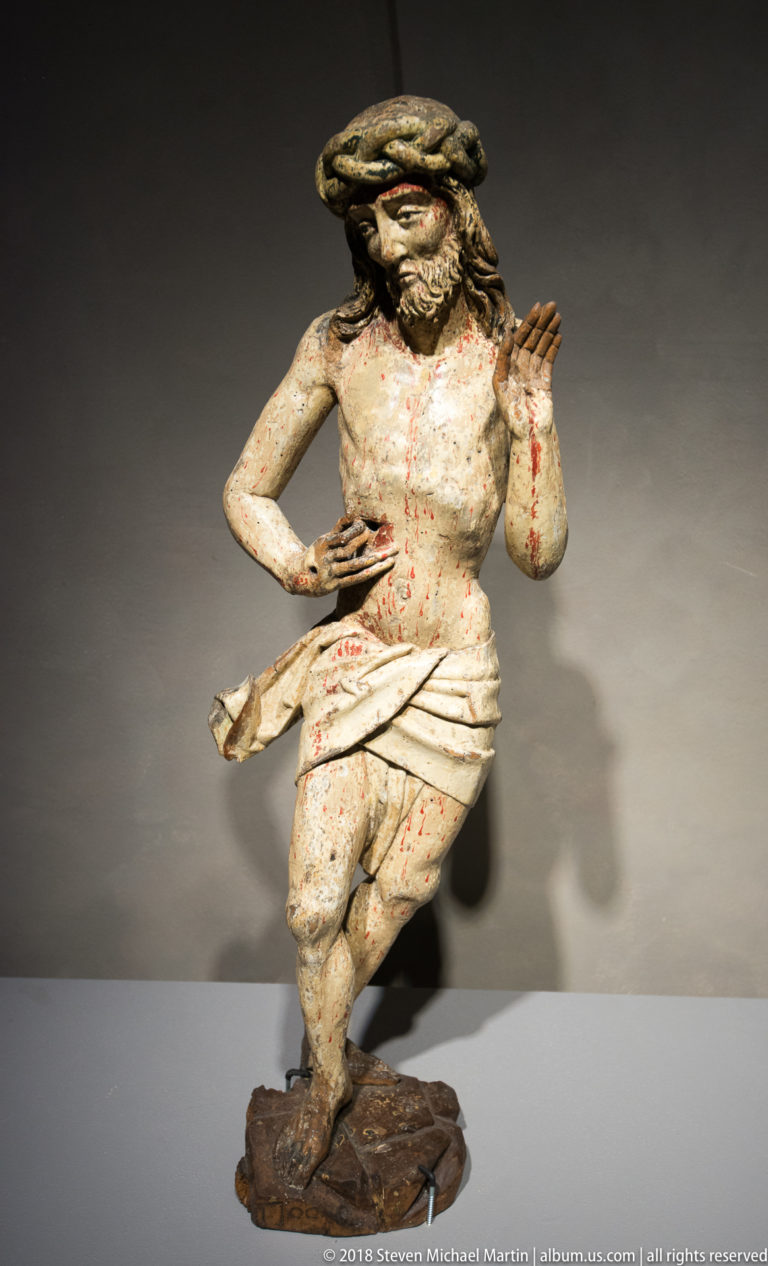 Polychrome carved Christ wearing a crown of thorns and loincloth, with painted wounds and one hand raised.