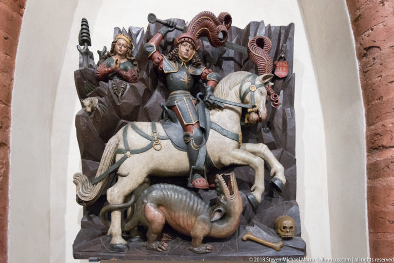 Polychrome relief of Saint George on a white horse attacking a dragon, with additional figures crowded into the background.