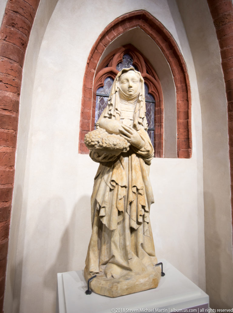 Carved pale stone female figure in a veil and layered robes, holding a shallow dish before a pointed window.