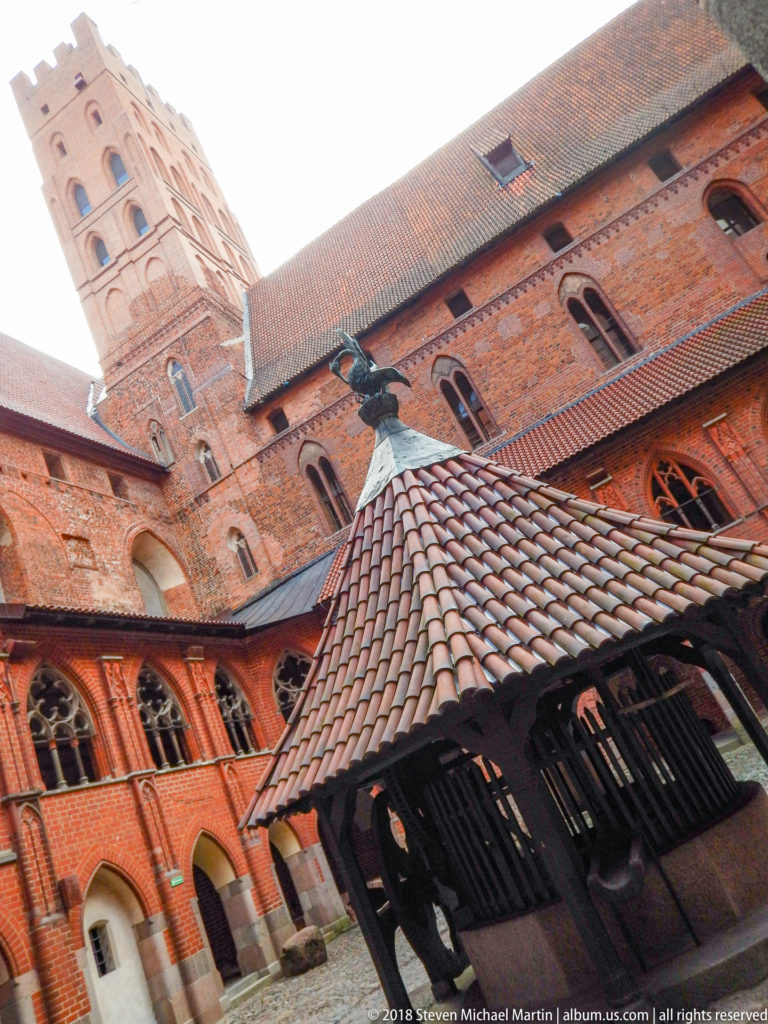 Low-angle view of a tiled roofed well beneath tall red-brick courtyard façades with Gothic windows and a tower above.