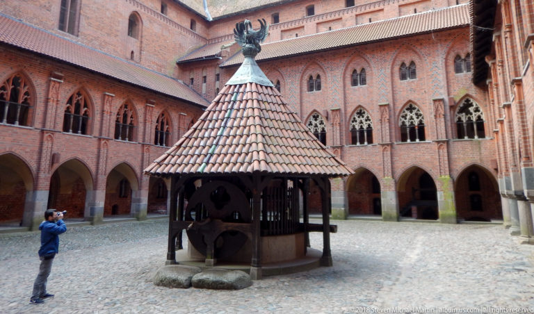 Visitor beside a roofed courtyard well, surrounded by red-brick arcades and tall pointed windows.