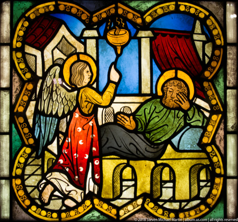 Winged angel beside a reclining haloed figure on a bed, with red drapery and blue architectural openings.