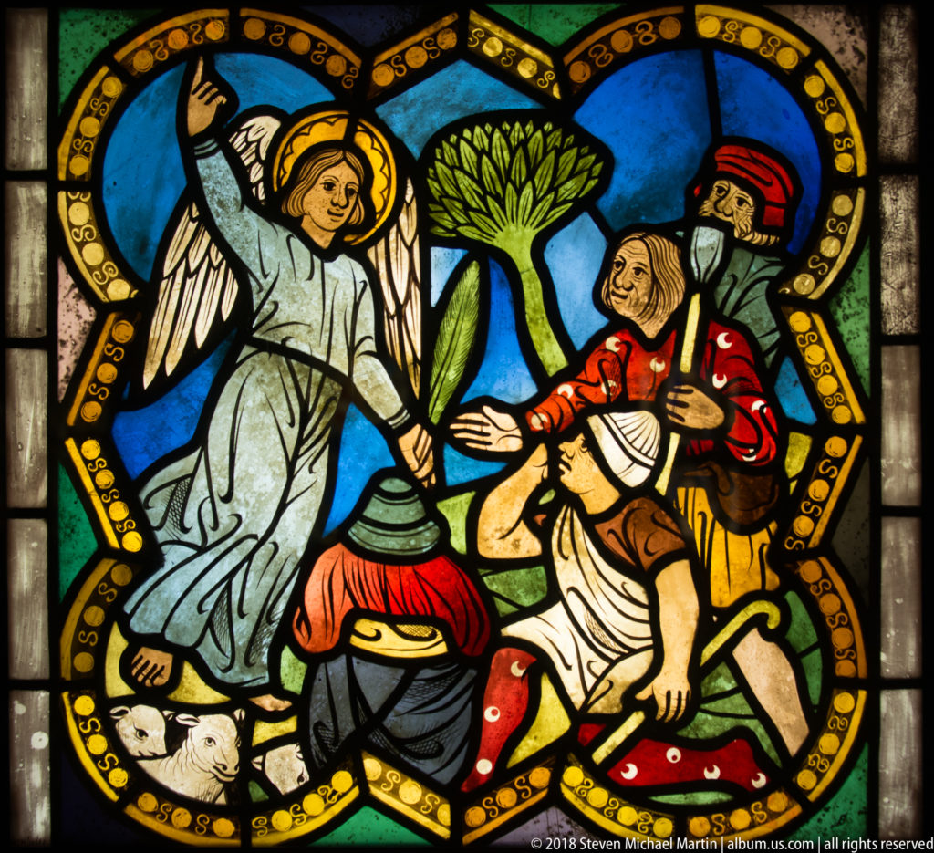 Winged angel facing two men beneath a large green tree in a medieval stained-glass landscape scene.
