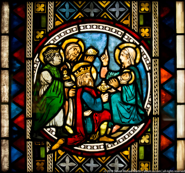 Crowned kneeling man before a haloed woman and child, with two attendants inside a round stained-glass medallion.
