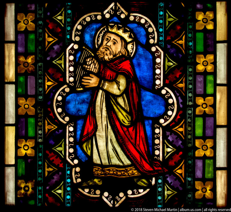Crowned haloed king in red mantle playing a small harp within a blue and multicolored stained-glass quatrefoil.