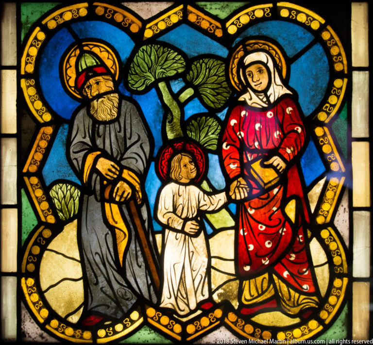Haloed bearded man, child, and woman walking together through a tree-filled medieval stained-glass landscape.