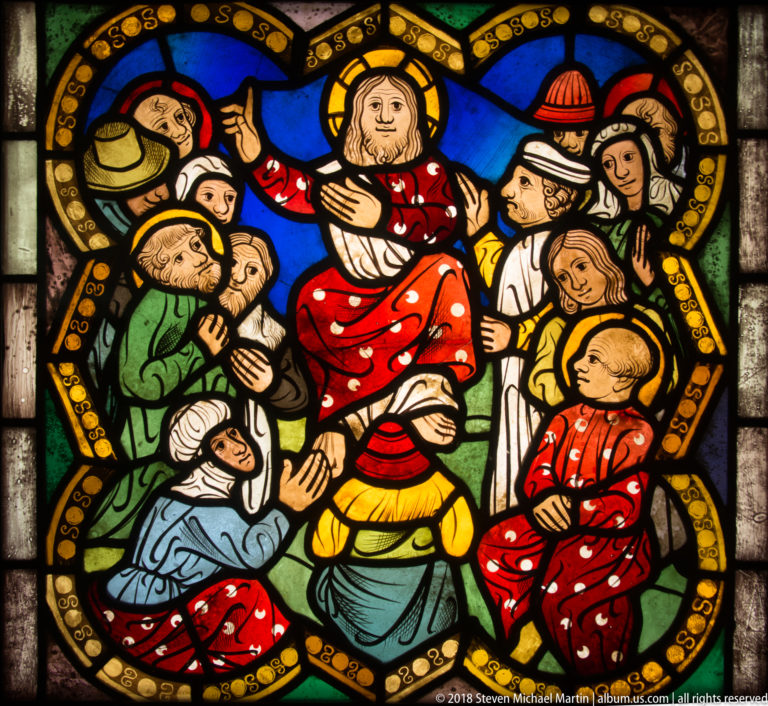 Haloed Christ in red robes addressing a tightly packed crowd of men and women in a stained-glass narrative scene.
