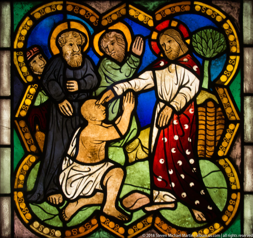 Christ reaches toward a kneeling man in a medieval stained-glass scene, with several onlookers framed by an ornamental border.