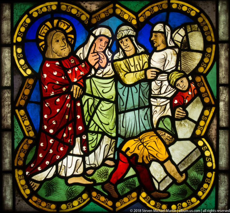 Haloed Christ in a red robe addresses several figures while one person bends low at the right of the stained-glass panel.