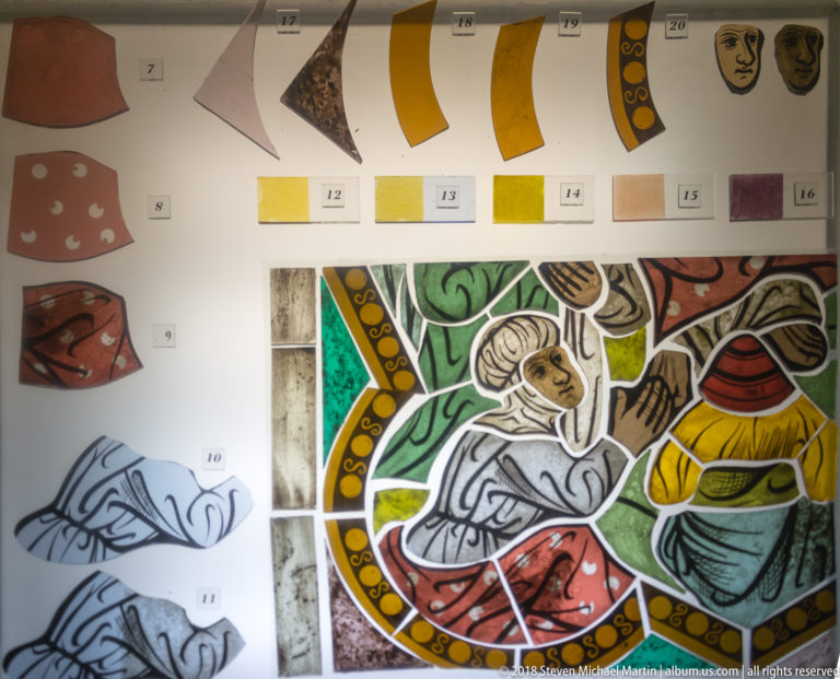 Technical stained-glass display with design studies, numbered color references, glass fragments, painted trials, and a reconstructed figural section.