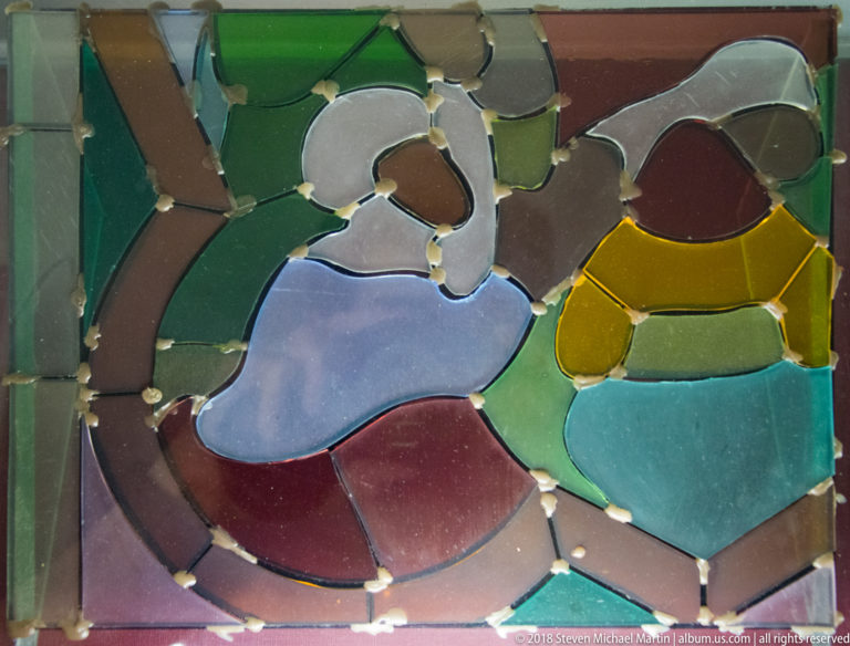 Individually cut green, violet, turquoise, yellow, and burgundy glass pieces fitted together to form part of a figure.