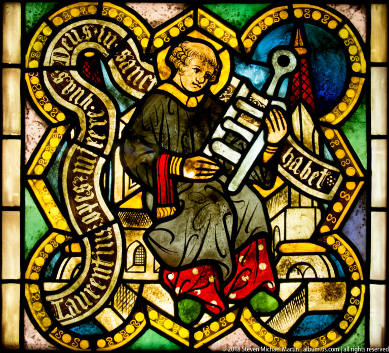 Saint Lawrence seated with his large gridiron attribute amid architectural forms and an inscribed ribbon.